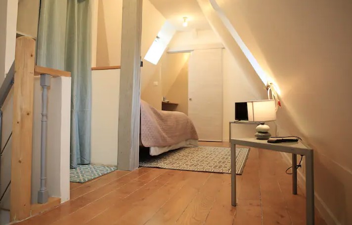 Chambre mansardée avec lit, table de nuit et éclairage doux. Sol en bois clair.